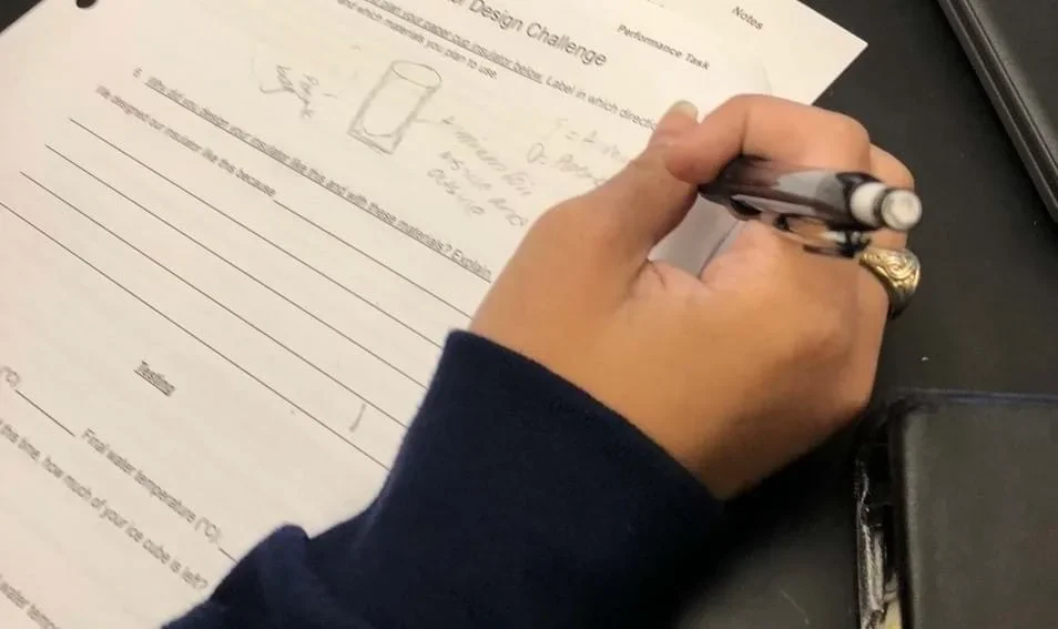 Close-up photo of a student’s right hand holding a pencil and writing on a piece of paper. The paper includes a drawing of a cup with labels around it.