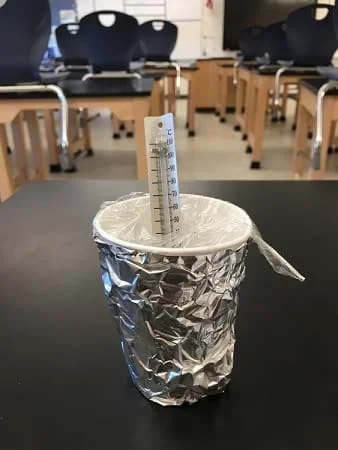 An insulated cup design filled with water sits on the table. The cup is covered in foil and has a lid of thin plastic wrap. A thermometer breaches the plastic lid and sits in the cup.