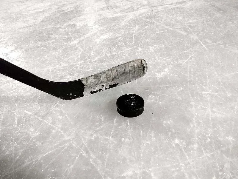 An image shows a hockey stick and hockey puck resting on a scratched ice rink.