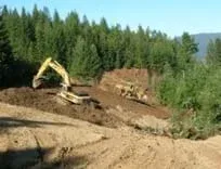 Two large excavators are removing dirt and trees along the side of forested mountain.