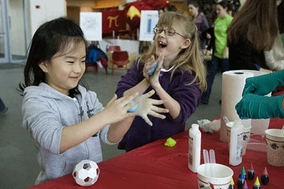 Photo shows two young girls laughing and smiling as they roll blue imitation silly putty in their hands.