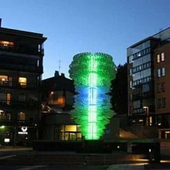 A photograph shows an illuminated green and blue (somewhat) tornado-shaped five-story light sculpture in a courtyard with seven-story buildings nearby.