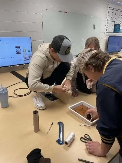 Three students stand around a table. A container with water mixed with dyed brown oil is on the table next to other materials. One student is skimming the top of the liquid with a cup.