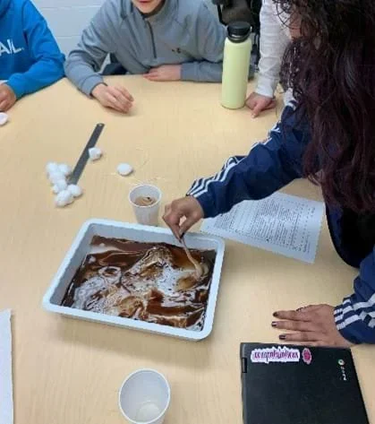 Oil dyed brown floats in water inside a container. The oil forms round bubbles to avoid mixing with the water as much as possible. A student uses a popsicle stick to carefully mix the dyed oil and the water.