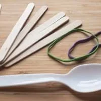 A close-up image of a plastic spoon, two rubber bands, and five popsicle sticks on a table.