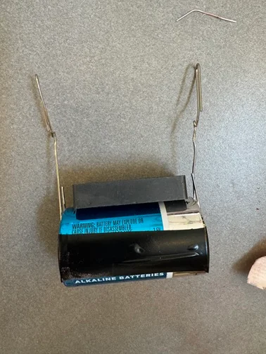 A photo of a D-cell battery with an unfolded paper clip taped with electrical tape to either end of the battery leads and a magnet placed on the battery side between the paper clips.