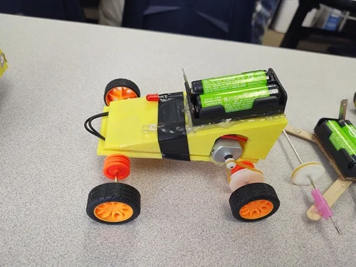 A photo of a mini EV design made of a 3D-printed triangular-shaped yellow body, plastic wheels, wire axles, plastic pulleys connected with a rubber band on the axle and motor spindle, which is powered by a motor and 2 AA battery pack.