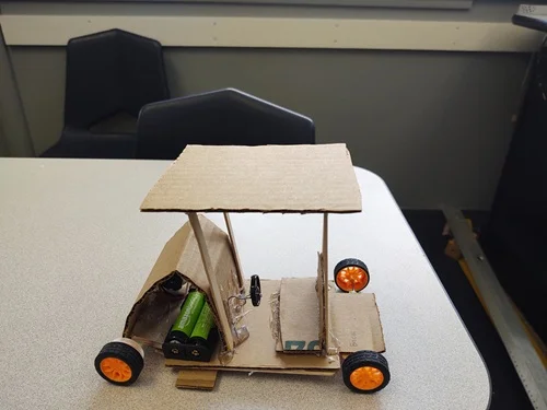 A photo of a mini EV design made with a cardboard chassis and car body that is powered by a motor and 2 AA battery pack.