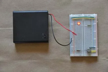Photo shows a breadboard (circuit board) with the following connections: a yellow wire, which connects the positive terminals on either side of the board; a green wire, which connects the negative terminals on either side of the board; an LED, which is connected to the positive terminal on one side, and the center of the board on the other side; a resistor, which is connected to the same column as the LED in the center of the board, and the negative terminal on the other side; a VOC sensor; a battery pack. The LED is illuminated.