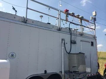 A photograph shows a white box-shaped trailer on wheels with railings around the rooftop onto which equipment is attached, including monitors, sensors, hoses and inlets into the trailer.