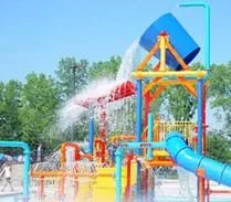 Photo shows an interactive play fountain featuring pressurized spouts of water and an elevated giant bucket with water pouring out