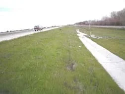 Photo shows a grassy area to the side of a highway, sloping down to a concrete collection drain.