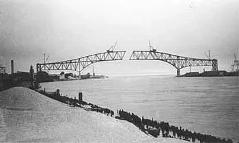 Black and white photo shows silhouette of two truss-type bridge spans being built from each side of a river, not yet meeting in the middle.