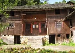 Photo shows the front view of a two-story wooden house raised on a stone foundation with a peaked tile roof, four windows and a centered double front door.
