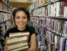 Photo shows a young girl holding a stack of books, surrounded by shelves full of books in a library.