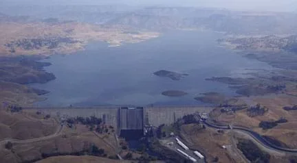 Aerial photo shows a wide and straight concrete structure blocking a river flow and holding back a supply of water. From above, the structure shape looks like a wall across the river width, tipped towards the resulting reservoir.