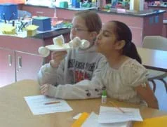 Photo of one girl holding a paper plate and paper cup contraption, and another girl blowing on it.