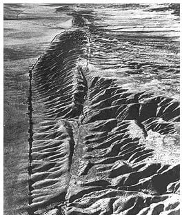 A black and white aerial photograph shows what looks like a slice through a ridge of desert mountains and foothils. It is the San Andreas fault in the Carrizo Plan in central California.