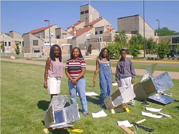 A photo shows four teens and three shiny, box-type solar ovens in a sunny field on a university campus.