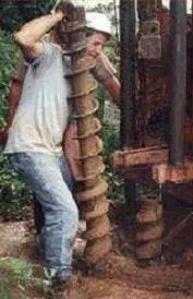 Photo shows a man drilling a hole in the ground with a drill (auger) that is as big as he is.
