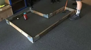 A photograph shows a wooden frame placed on a carpeted floor. The frame edges are made of 2 x 4-inch wood with galvanized metal corner joints as connectors and enclose an L-shaped interior space. A student has just used a golf putter to hit a golf ball from one end of the L shape towards a water bottle (which serves as the hole/goal) in the far side of the L shape.