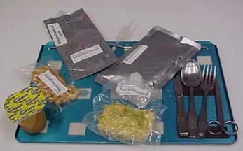 Photo shows a blue tray with a metal knife, spoon, fork and scissors; two labeled foil-sealed packages of food or drink; two clear plastic vacuum-sealed packages of food, and a pudding cup. The packages are attached to the tray with Velcro.