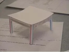 A "finished" model parking garage with four posts and a corrugated top. The posts are made of plastic drinking straws and the top is made of two layers of white poster board with a corrugated center.