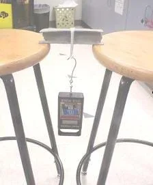 This photograph shows a seven-inch clay I-beam sitting on the edges of two stool seats, spanning a six-inch gap between the two stools, with a weight hanging from a loop of string placed around the middle of the beam.