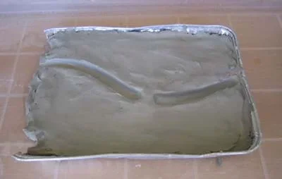 Baking pan filled with clay shaped into a slightly curved riverbed with a break at one point in the riverbed.