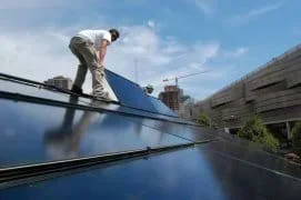 Two men standing on an angled roof lay a shiny blue-black panel on its surface.