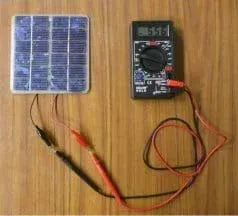 Photo from above a tabletop shows red and black wires from a small PV panel connected to red and black leads from a digital multimeter.