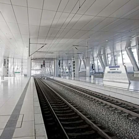 A photo shows the interior tunnel of a modern railway station with two sets of train tracks, ceiling, windows and waiting platforms.