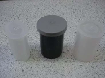 A photograph shows three types of film canisters sitting on a counter top: (left) a translucent plastic container and lid that are elliptical in shape; (center) a 35 mm photographic film canister made of black plastic with a gray overlapping cap; (right) a 35 mm photographic film canister made of translucent plastic with an inset fitted cap.