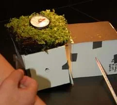 Photo shows two small cube structures made of taped-together foam core board. One rooftop has a square of sod with a round thermometer sitting on it. A long thermometer pokes into the interior of the other structure.
