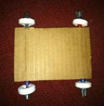 A photograph shows a view from above of two plastic coffee stirrers run through the cardboard layers of a 3 x 5 piece of corrugated cardboard, making axles in a car chassis base. Where the stirrers extend outside of the cardboard at four spots, wheels are attached by placement of a plastic bead and then ring-shaped hard candy, with the stirrer ends duct-taped.