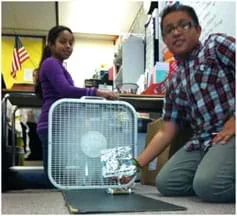 A photograph in a classroom shows a boy positioning a toy-sized sail car in front of a box fan while a girl operates the fan control switch.