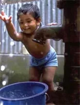 Photo shows a boy splashing water from a bucket under a steel water pump.