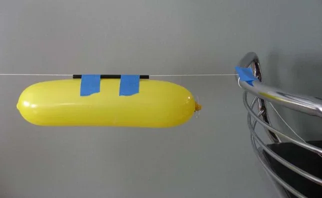 Photo shows an inflated long balloon taped to a drinking straw that hangs from a taut string attached to the back of a chair.