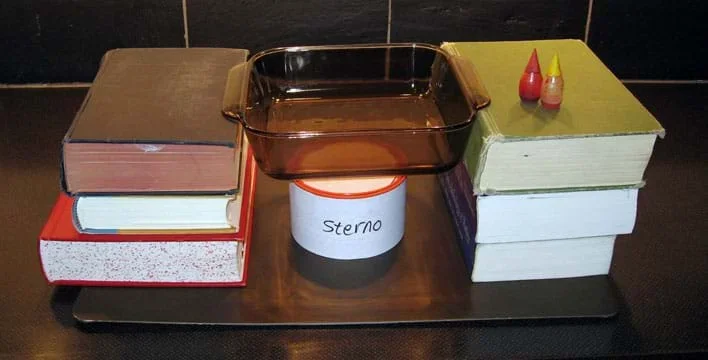 A glass baking dish is suspended by stacks of three books on each side over a can of sterno. Food coloring bottles are nearby.