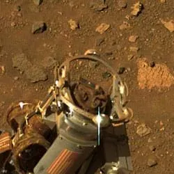 Photo shows a circular arrangement of moving bits at the end of a metal tube, against a background of a flat pavement of reddish brown soil and flat-surfaced angular rocks.