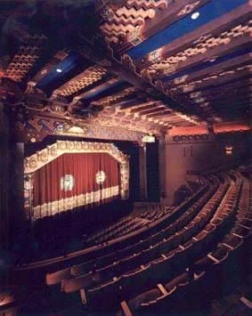 A photograph of the KiMo Theatre in Albuquerque, NM. Shown are rows of curved seats, each row rising above the previous row, a stage with a red curtain and corrugated panels on the ceiling.