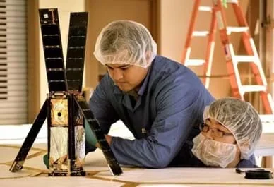 Two men wearing hair and beard nets examine a device on a table. Four solar panel-covered sides of the tall rectangular object (shoe-box sized) are flipped up or away to reveal the inside volume of the satellite cube, which contains a folded-up silver and gold fabric.
