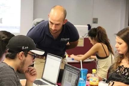 A photograph shows an instructor helping students as they work on computers.