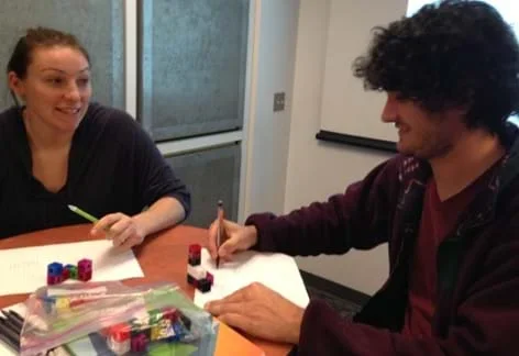 A photograph shows two teens at a table drawing two-dimensional illustrations on paper of the three-dimensional cubes on the table in front of them.