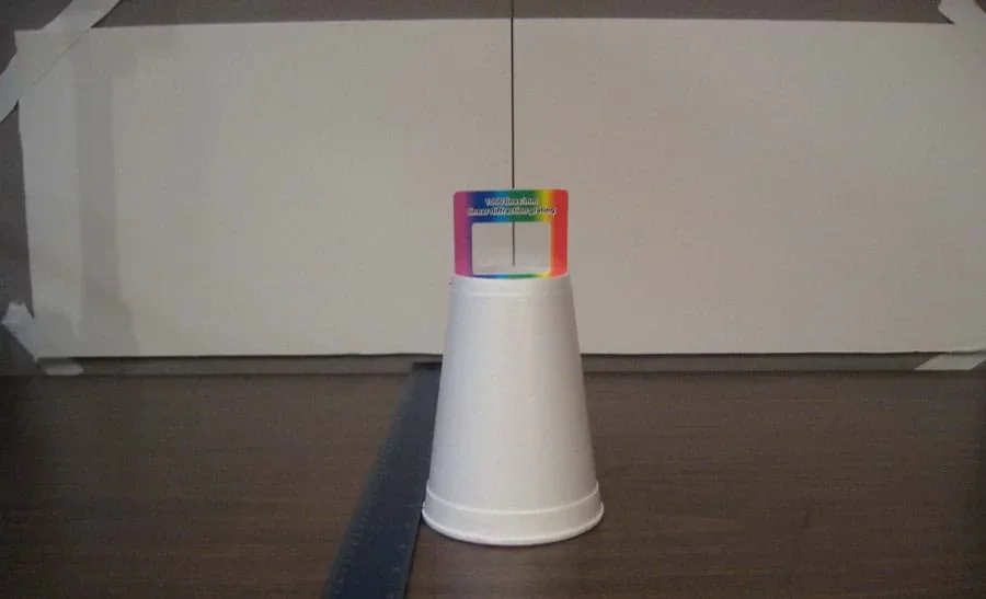 Photo shows an upside-down cup on a tabletop, lined up in front of a black vertical line in the middle of a wide white paper taped to the wall behind, with a ruler positioned on the table nearby.