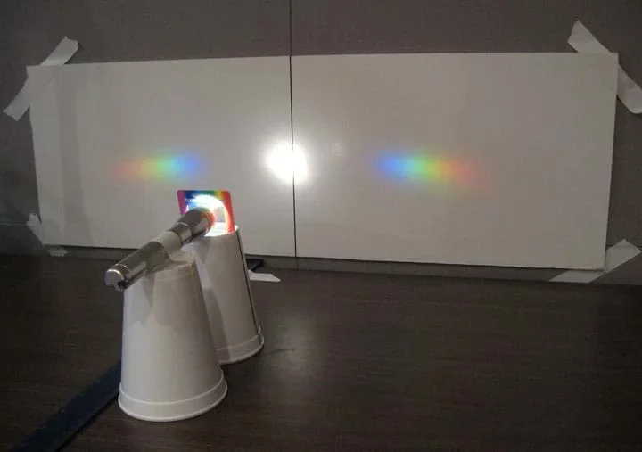 Photo shows a light shining through the grating, forming two rainbow-colored spectra to either side of a round, white light image in the center of the screen.
