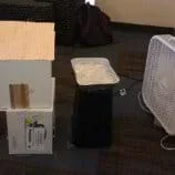 A photograph shows a small-sized foamcore board model house resting on an overturned cardboard box. On the right, a box fan blows air over a tray of ice toward the model house.