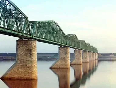 A photo shows a steel truss railroad bridge supported by eight brick pillars spans a still body of water. The truss design uses repeated triangular shapes composed of steel beams.