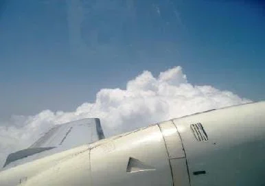 Photo from the window of an airplane shows airplane wing, blue sky and a patch of clouds.