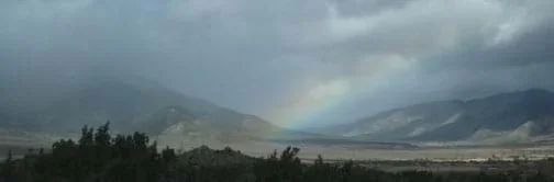 Landscape photo of stormy skies and a partial rainbow over a wide valley and mountain backdrop.
